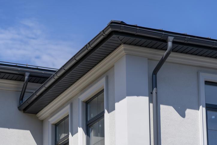 Corner of house with windows, new gray metal tile roof and rain gutter. Metallic Guttering System, Guttering and Drainage Pipe Exterior